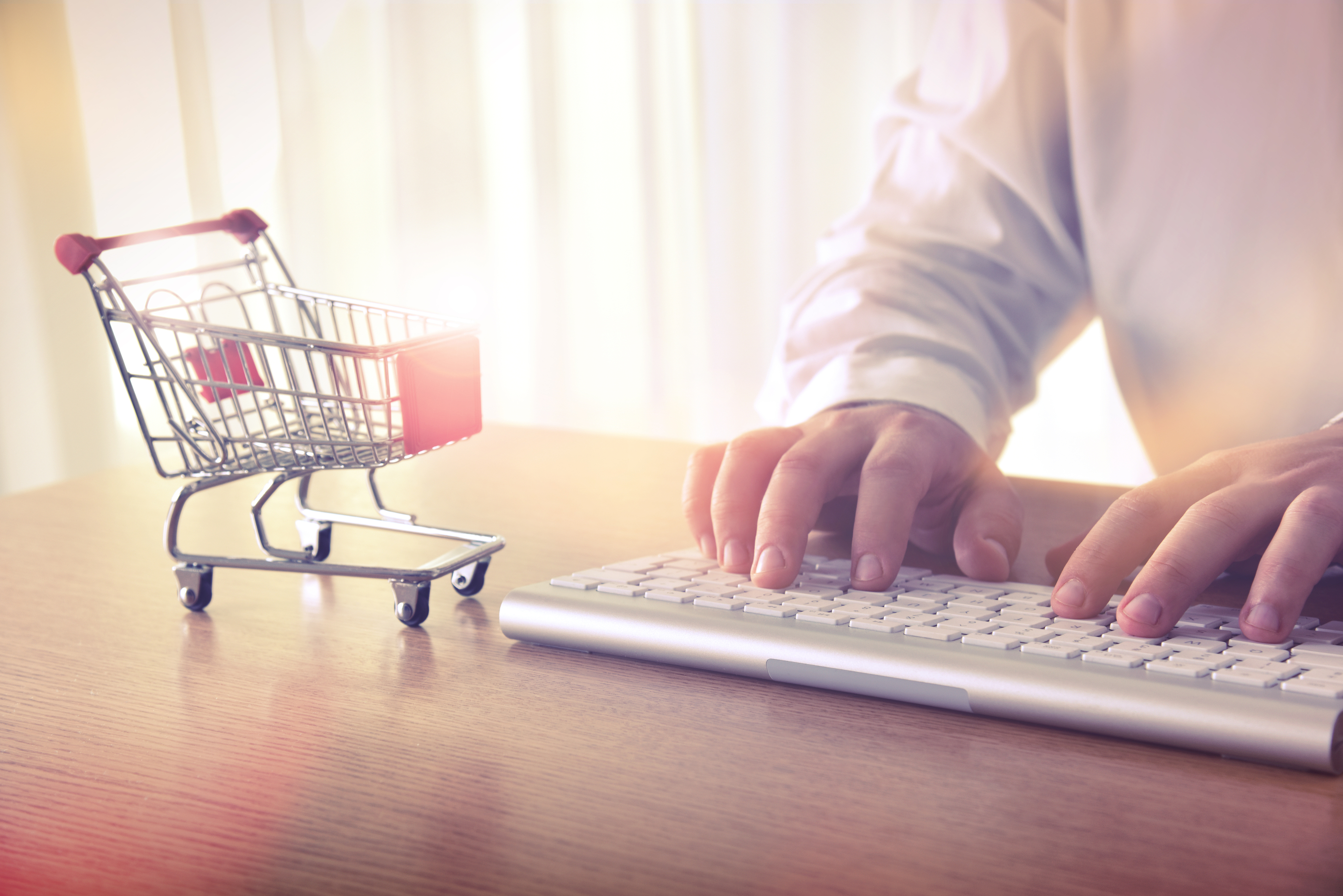 E-commerce concept. Man's hands typing on computer keyboard next to a shopping cart. Concept of online shopping and e-commerce, for background, website banner, promotional materials, presentation templates, advertising.